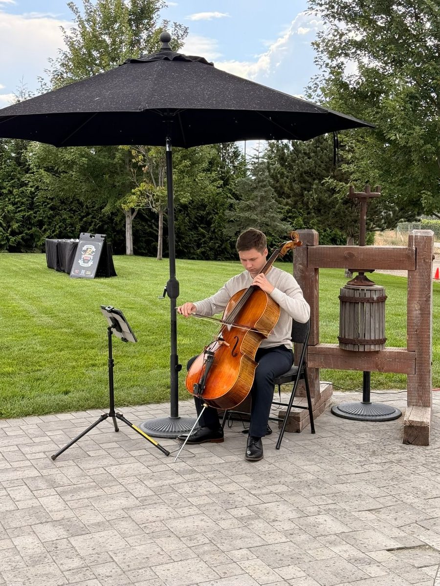 Yaroslav playing the cello outdoors at a wedding, shaded by a parasol